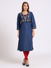 A woman modeling a blue denim-style tunic dress with colorful embroidered detailing around the neckline and sleeve cuffs, paired with red tights and navy heels, against a plain light gray studio background.