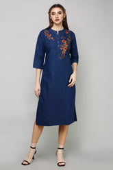 A woman modeling a dark blue cotton kurta with orange and red floral embroidery on the chest, paired with beige leggings and black sandals, against a solid gray studio background.