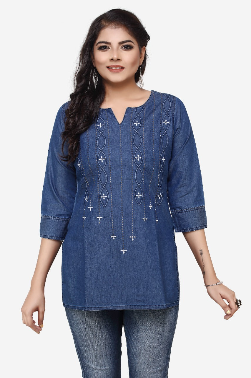 A woman modeling a blue denim tunic top with white embroidered details, paired with blue jeans, against a plain light gray background.