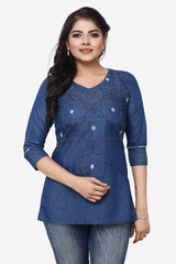 A woman modeling a blue denim long-sleeve tunic top with geometric embroidery and diamond-shaped appliques, paired with blue jeans against a light gray background.