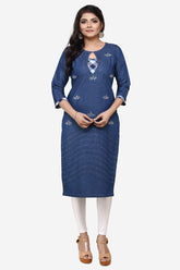A woman modeling a blue denim-style kurta with small embroidered details, paired with white leggings and brown sandals, against a plain light gray background.