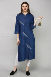 A woman modeling a blue denim-style tunic with white embroidered diagonal stripes, paired with white leggings and black slip-on shoes, against a plain gray studio background.