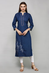 A woman modeling a dark blue denim-style kurta with white embroidery on the chest and lower skirt, paired with white leggings and tan heeled sandals, against a solid gray studio background.