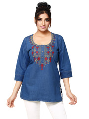 A woman modeling a blue denim tunic top with red and gold embroidered detailing around the neckline, paired with white pants, against a plain white background.