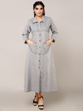 A woman modeling a light gray denim maxi shirt dress with a button-down front, collar, and front pockets, standing against a solid beige studio background.