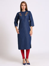 A woman modeling a dark blue denim-style kurta with colorful floral embroidery on the chest, paired with red leggings and navy blue heels, against a plain light gray studio background.