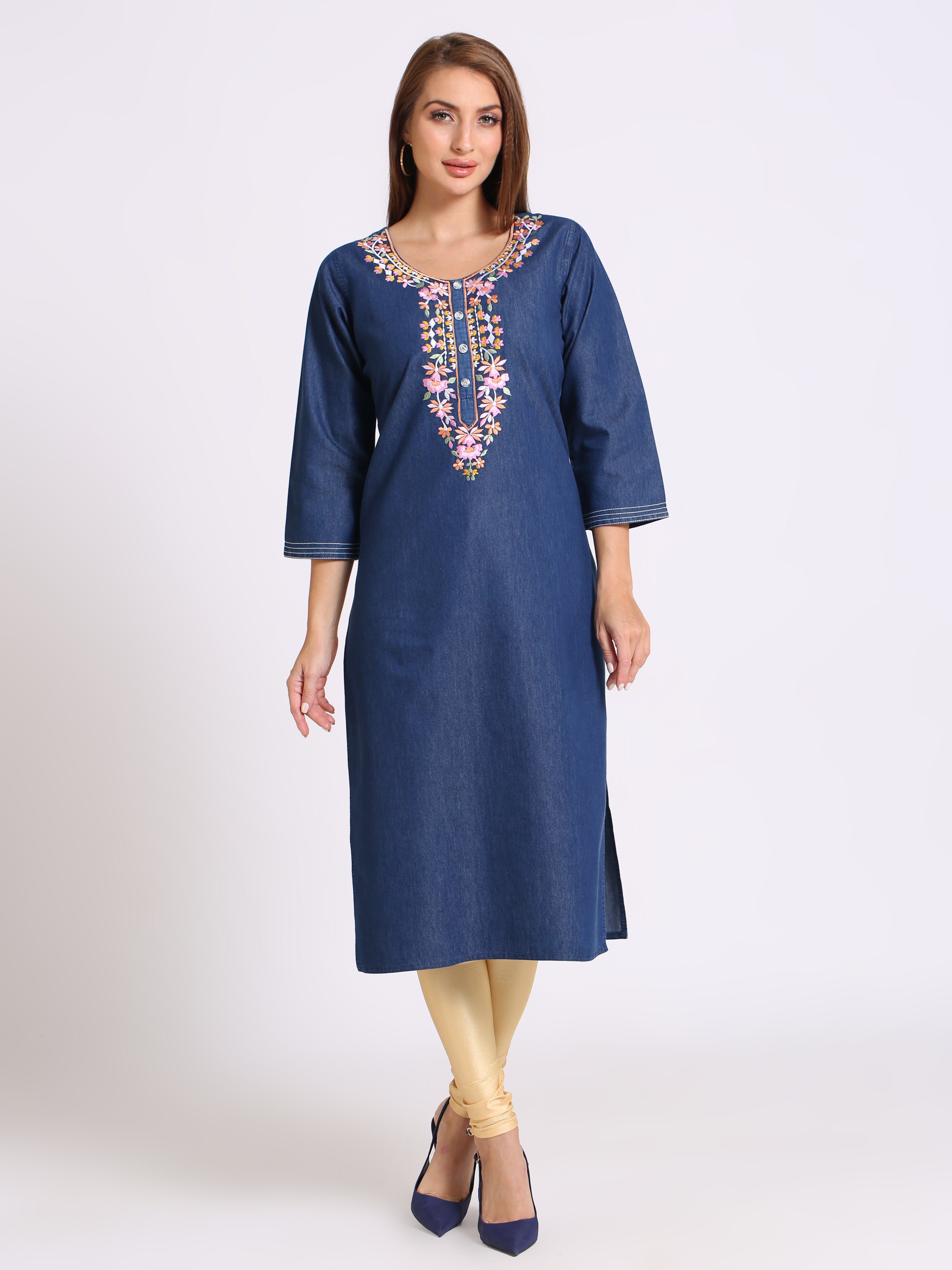 A woman modeling a blue denim tunic dress with colorful floral embroidery around the neckline, paired with beige leggings and navy blue heels, against a plain light gray studio background.