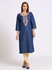 A woman modeling a blue denim tunic dress with colorful floral embroidery around the neckline, paired with beige leggings and navy blue heels, against a plain light gray studio background.