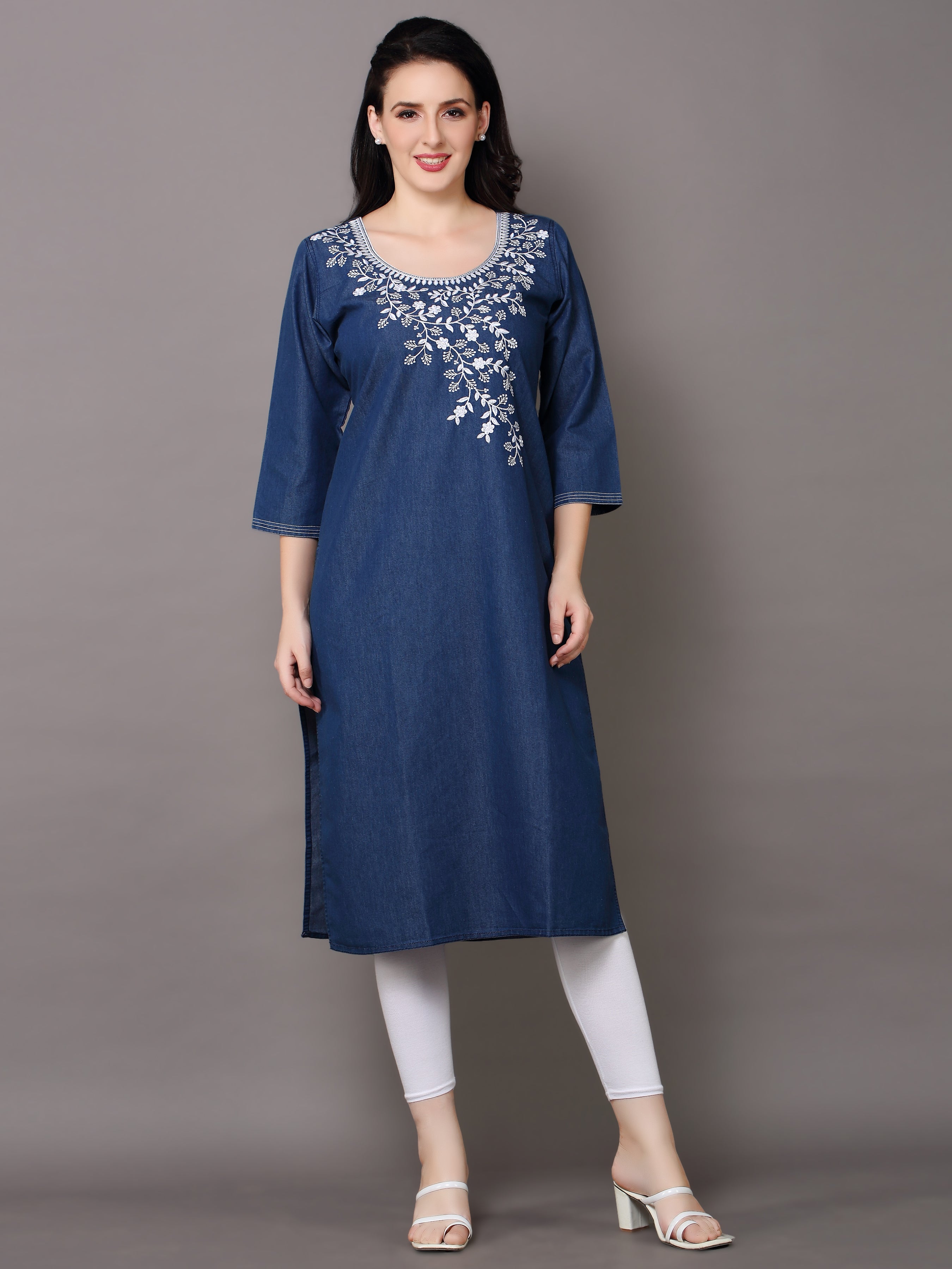 A woman modeling a blue denim-style A-line kurta with white floral embroidery around the neckline, paired with white leggings and white heeled sandals, against a solid gray studio background.