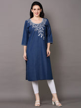A woman modeling a blue denim-style A-line kurta with white floral embroidery around the neckline, paired with white leggings and white heeled sandals, against a solid gray studio background.