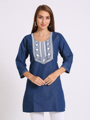 A woman modeling a blue denim tunic with white embroidered detailing around the neckline, set against a plain light gray background.