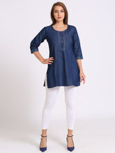 A woman modeling a blue denim tunic top with three-quarter sleeves, white leggings, and navy blue heels against a plain white studio background.