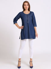 A woman modeling a blue denim tunic top with three-quarter sleeves, white leggings, and navy blue heels against a plain white studio background.
