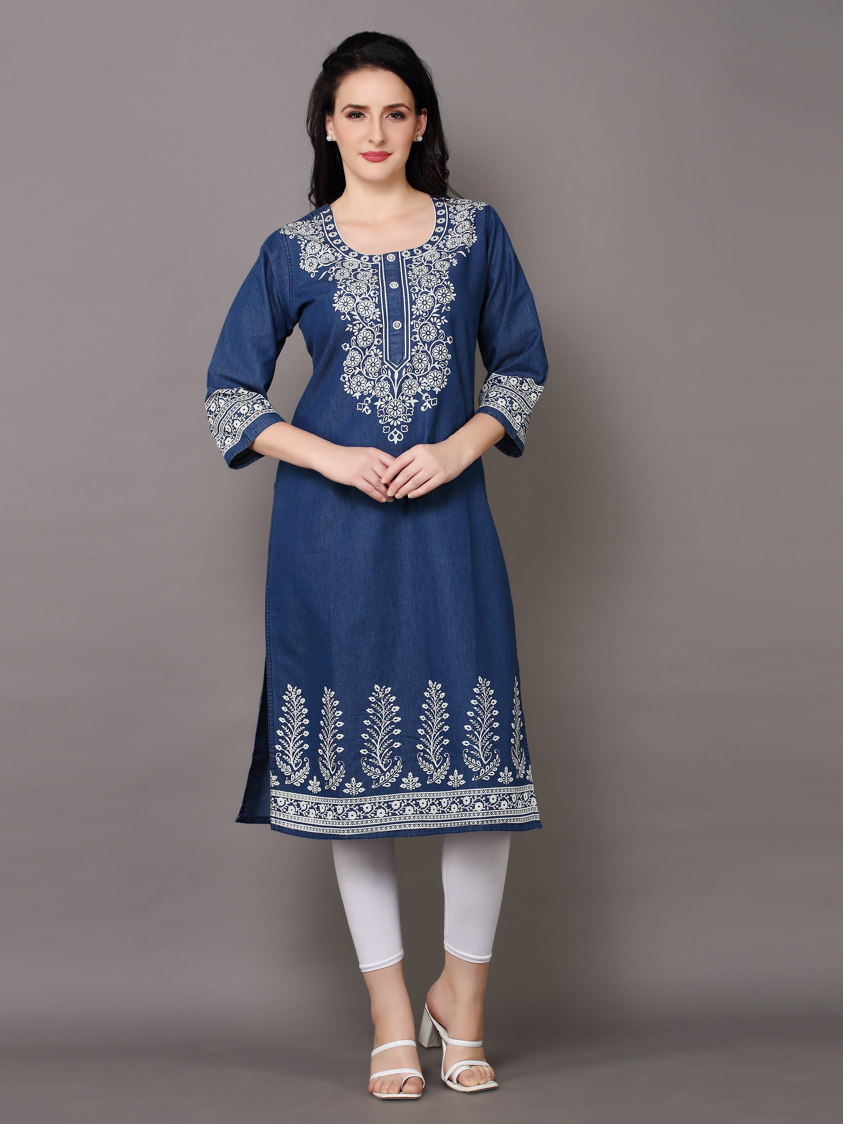 A woman modeling a navy blue kurta with intricate white embroidery on the neckline, sleeves, and hem, paired with white leggings and white heeled sandals against a solid gray studio background.