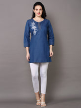 A woman modeling a blue cotton kurta with white floral embroidery on the chest, paired with white leggings and beige sandals, against a solid gray studio background.