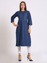 A woman modeling a dark blue denim-style tunic dress with white embroidered details on the chest, paired with white leggings and navy blue heels, against a plain light gray studio background.