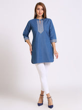 A woman modeling a blue cotton kurta with white printed embroidery on the chest and cuffs, paired with white pants and blue heels, against a light gray studio background.