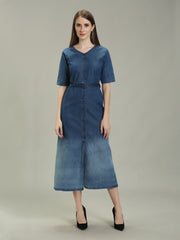A woman modeling a short-sleeved blue denim midi dress with a button-down front and a matching waist tie, paired with black heels, against a neutral gray studio background.