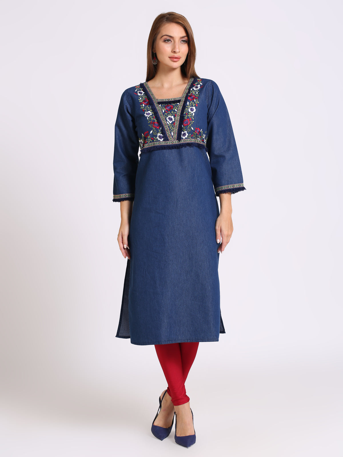 A woman modeling a blue denim-style tunic dress with colorful embroidered detailing around the neckline and sleeve cuffs, paired with red tights and navy heels, against a plain light gray studio background.