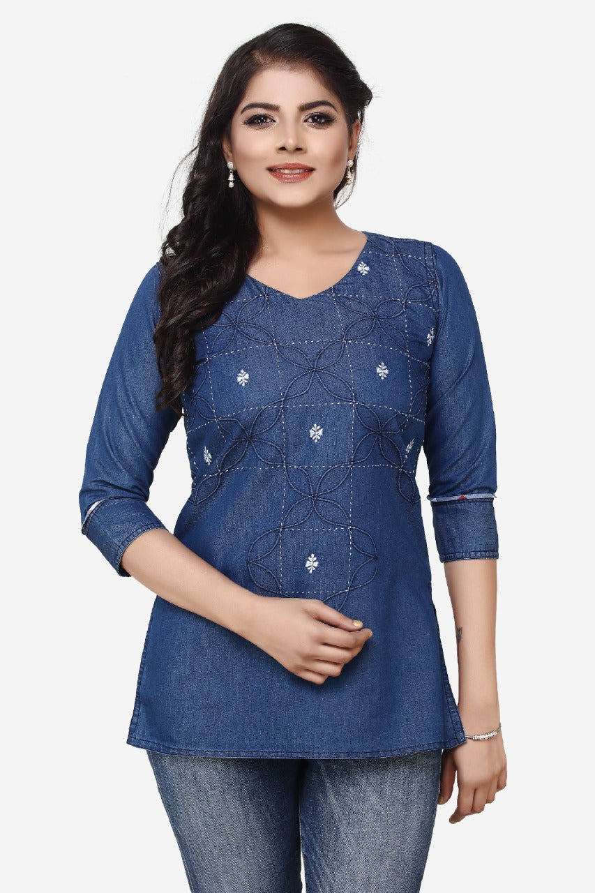 A woman modeling a blue denim long-sleeve tunic top with geometric embroidery and diamond-shaped appliques, paired with blue jeans against a light gray background.