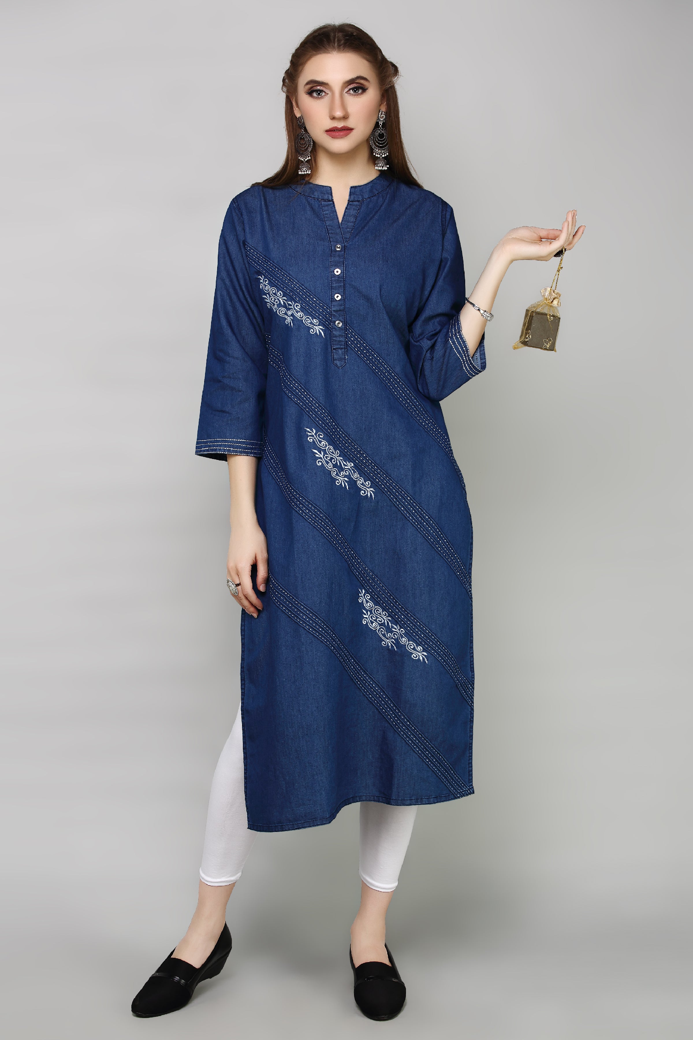 A woman modeling a blue denim-style tunic with white embroidered diagonal stripes, paired with white leggings and black slip-on shoes, against a plain gray studio background.
