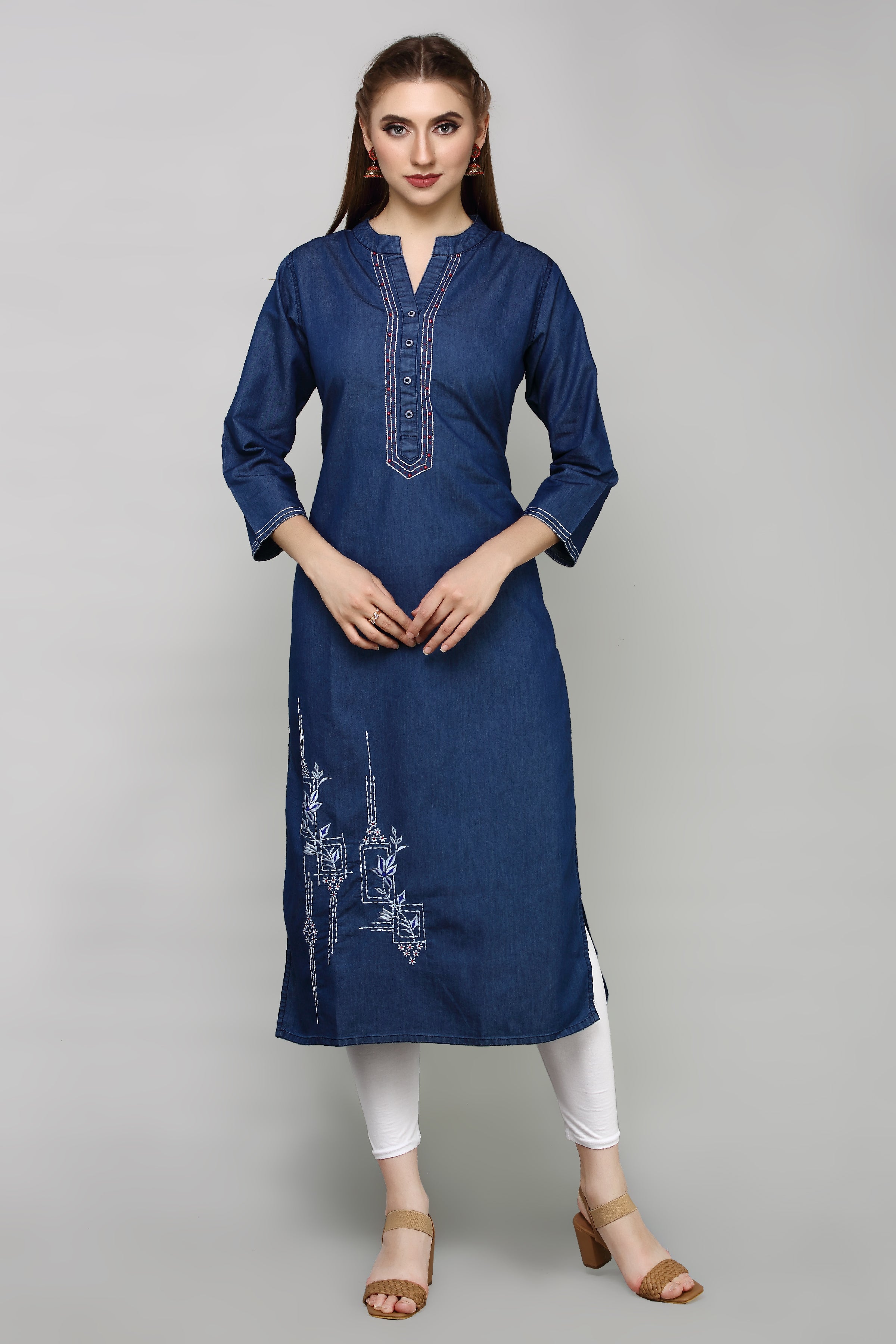 A woman modeling a dark blue denim-style kurta with white embroidery on the chest and lower skirt, paired with white leggings and tan heeled sandals, against a solid gray studio background.