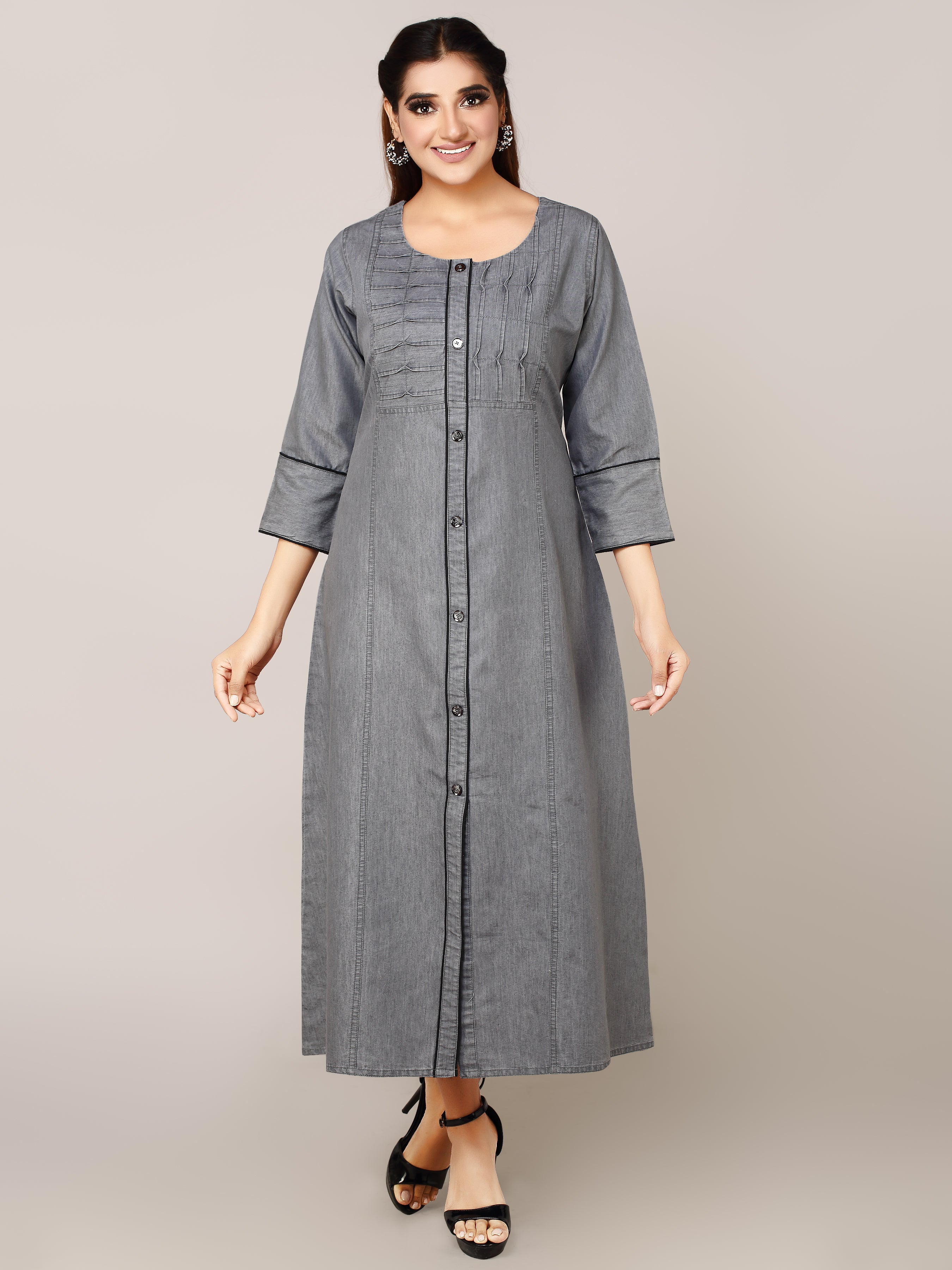 A woman modeling a long gray button-front tunic dress with three-quarter sleeves and subtle vertical pleating details, photographed against a plain light gray studio background.