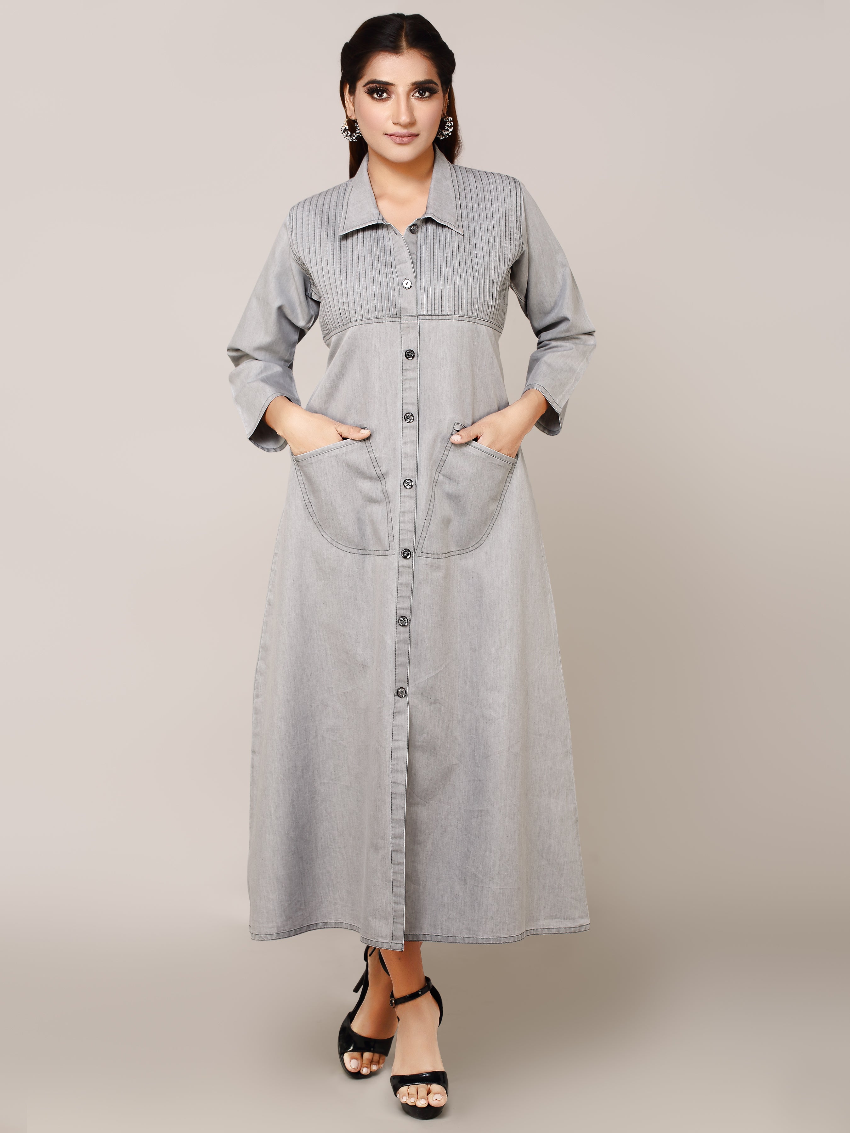 A woman modeling a light gray denim maxi shirt dress with a button-down front, collar, and front pockets, standing against a solid beige studio background.