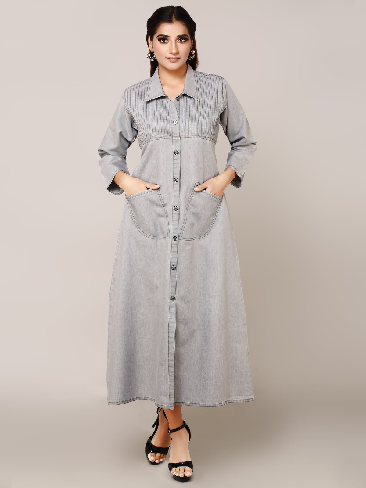 A woman modeling a light gray denim maxi shirt dress with a button-down front, collar, and front pockets, standing against a solid beige studio background.