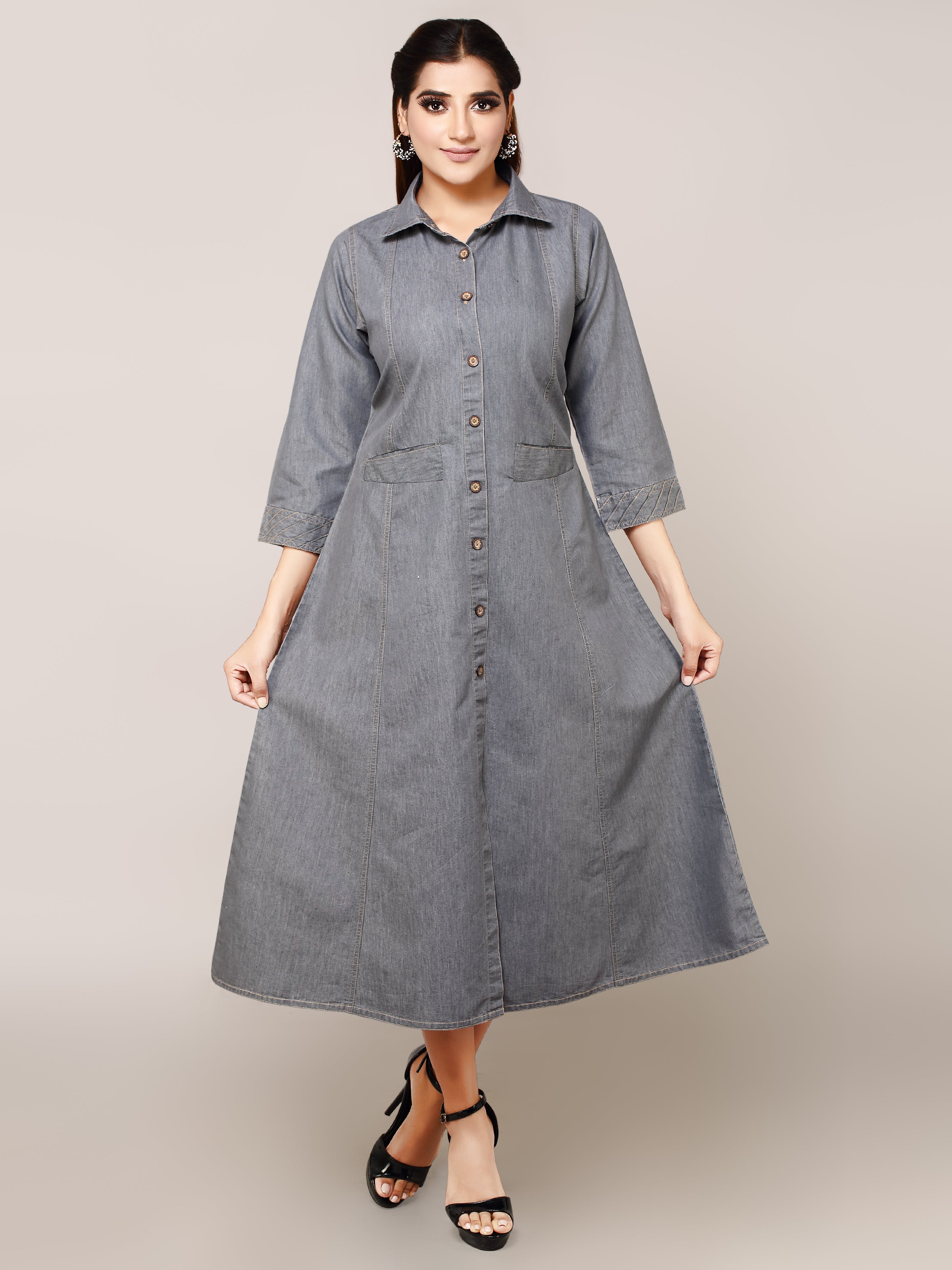 A woman modeling a gray denim midi shirt dress with three-quarter sleeves, a button-down front, and side pockets, paired with black strappy heels against a solid beige studio background.