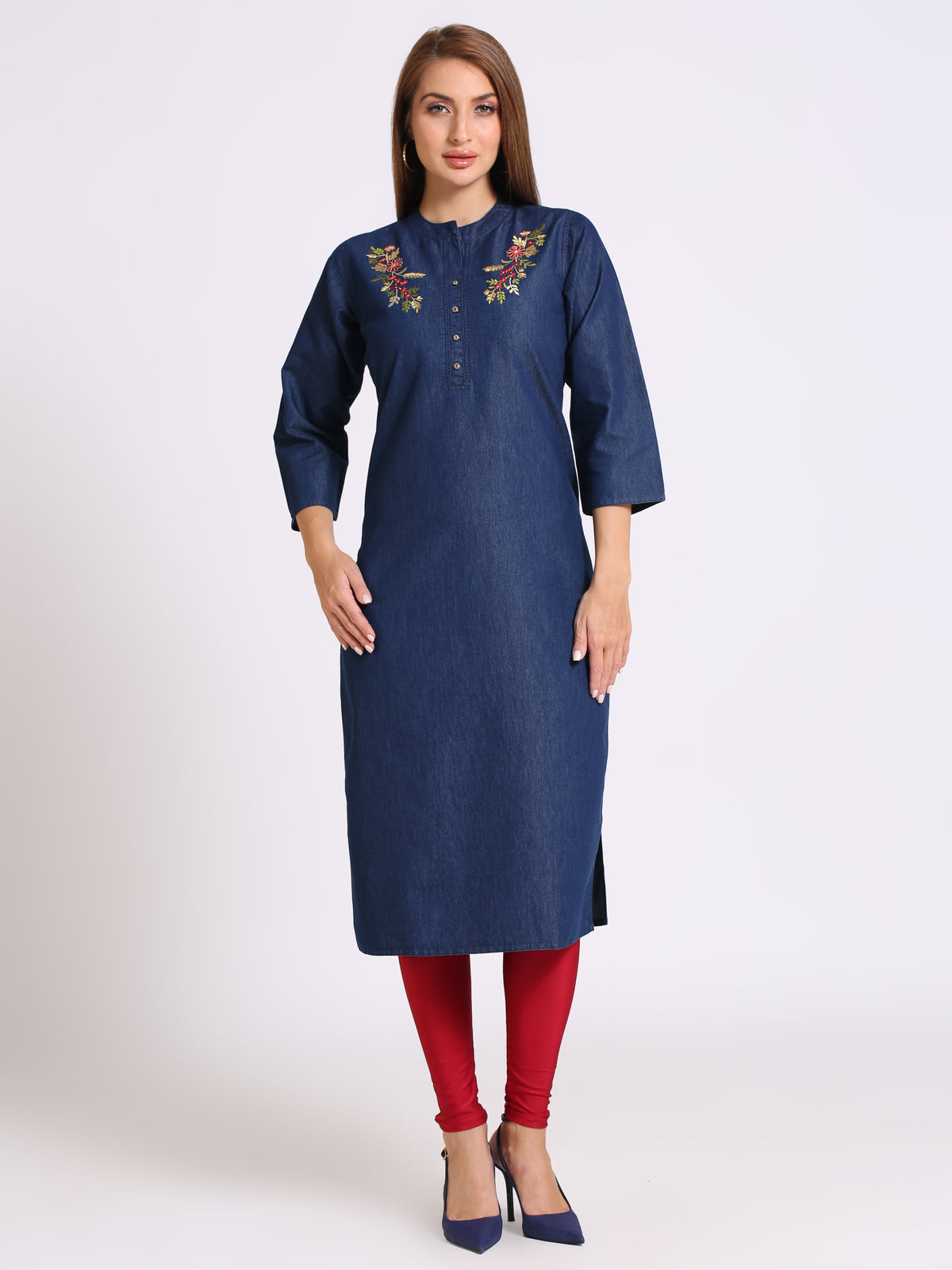 A woman modeling a dark blue denim-style kurta with colorful floral embroidery on the chest, paired with red leggings and navy blue heels, against a plain light gray studio background.