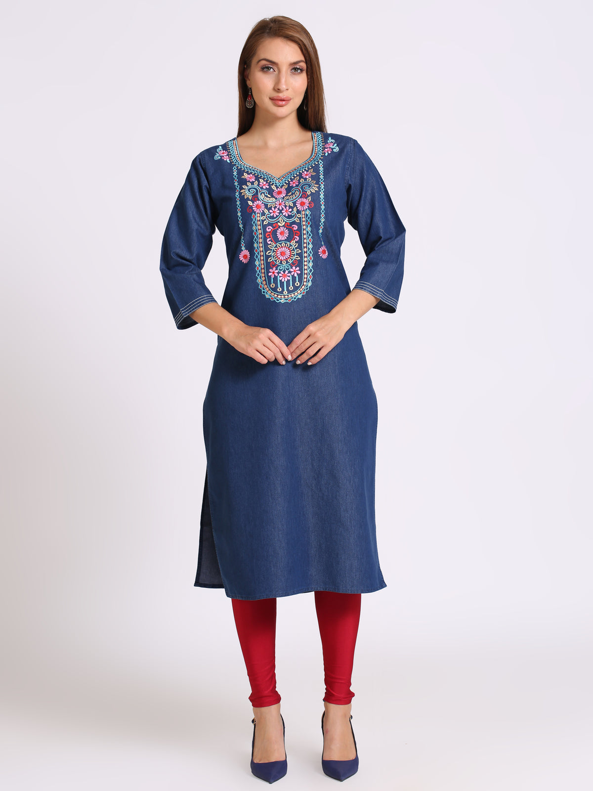 A woman modeling a blue denim-style kurta with colorful floral embroidery around the neckline, paired with red leggings and navy heels, against a plain light gray studio background.