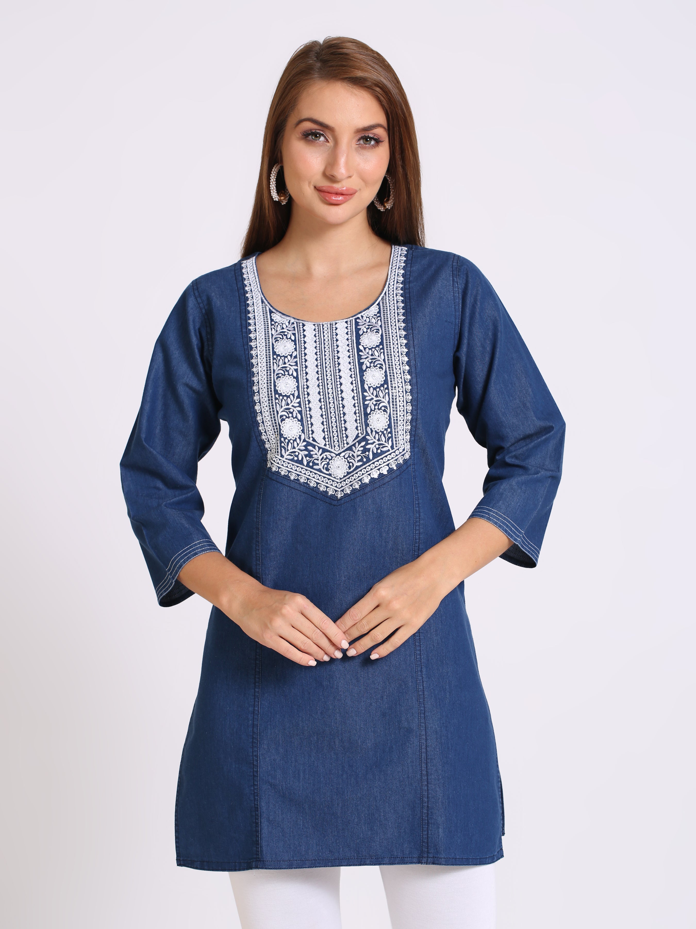 A woman modeling a blue denim tunic with white embroidered detailing around the neckline, set against a plain light gray background.