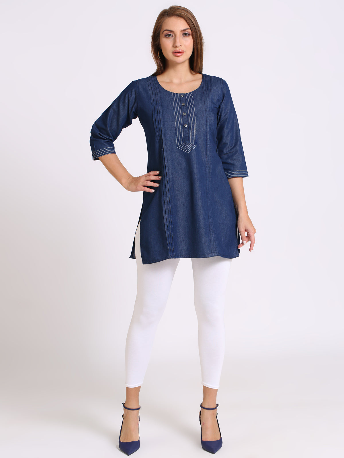 A woman modeling a blue denim tunic top with three-quarter sleeves, white leggings, and navy blue heels against a plain white studio background.