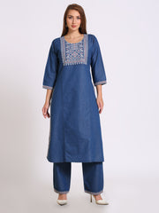 A woman modeling a blue three-piece Indian ethnic wear set, consisting of a long-sleeved kurta with white embroidery around the neckline and sleeve cuffs, matching wide-leg trousers, and white heels, set against a plain light gray background.