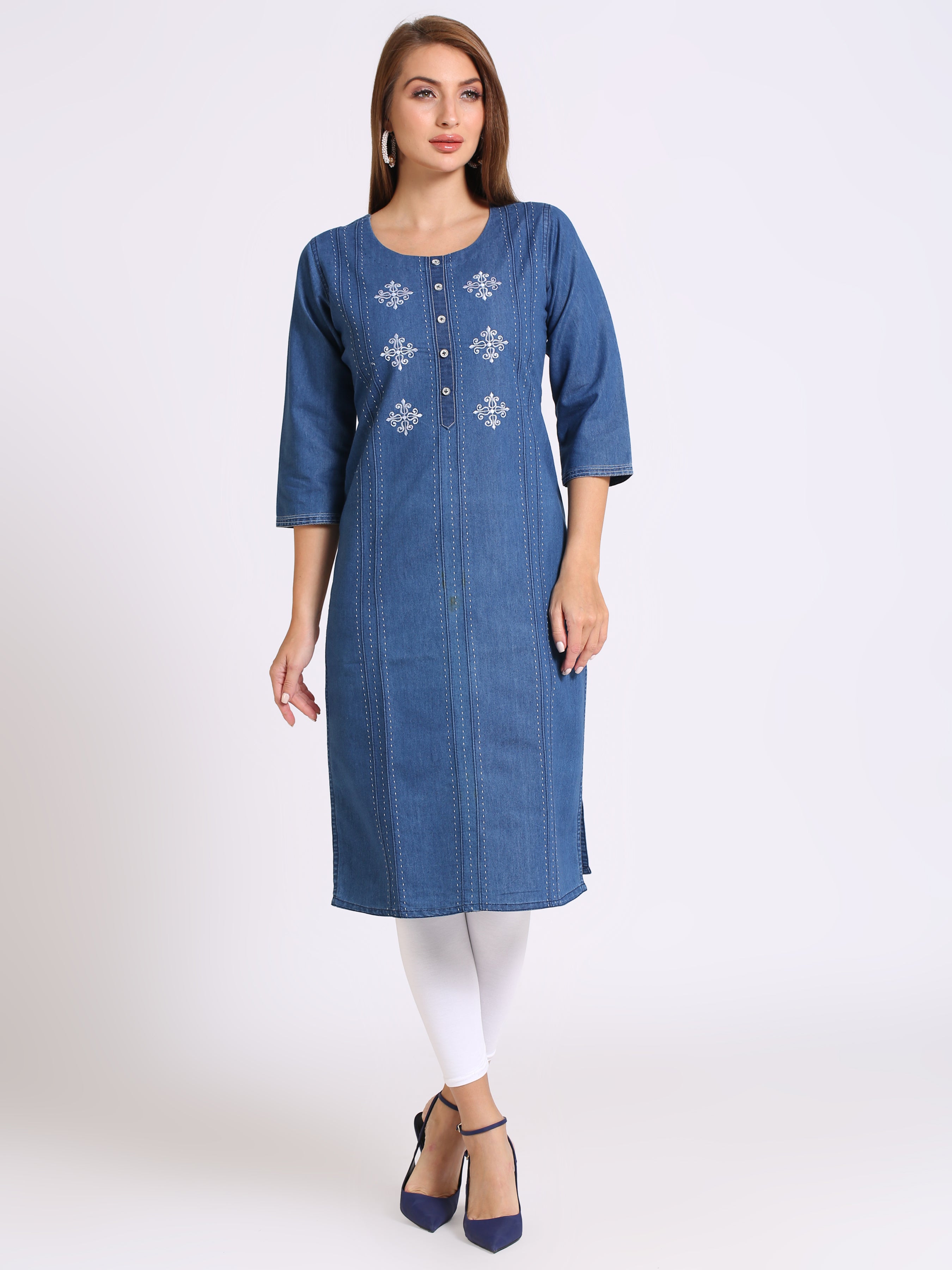 A woman modeling a blue denim-style tunic with white embroidered floral details on the chest, paired with white leggings and navy heels, against a plain light gray studio background.