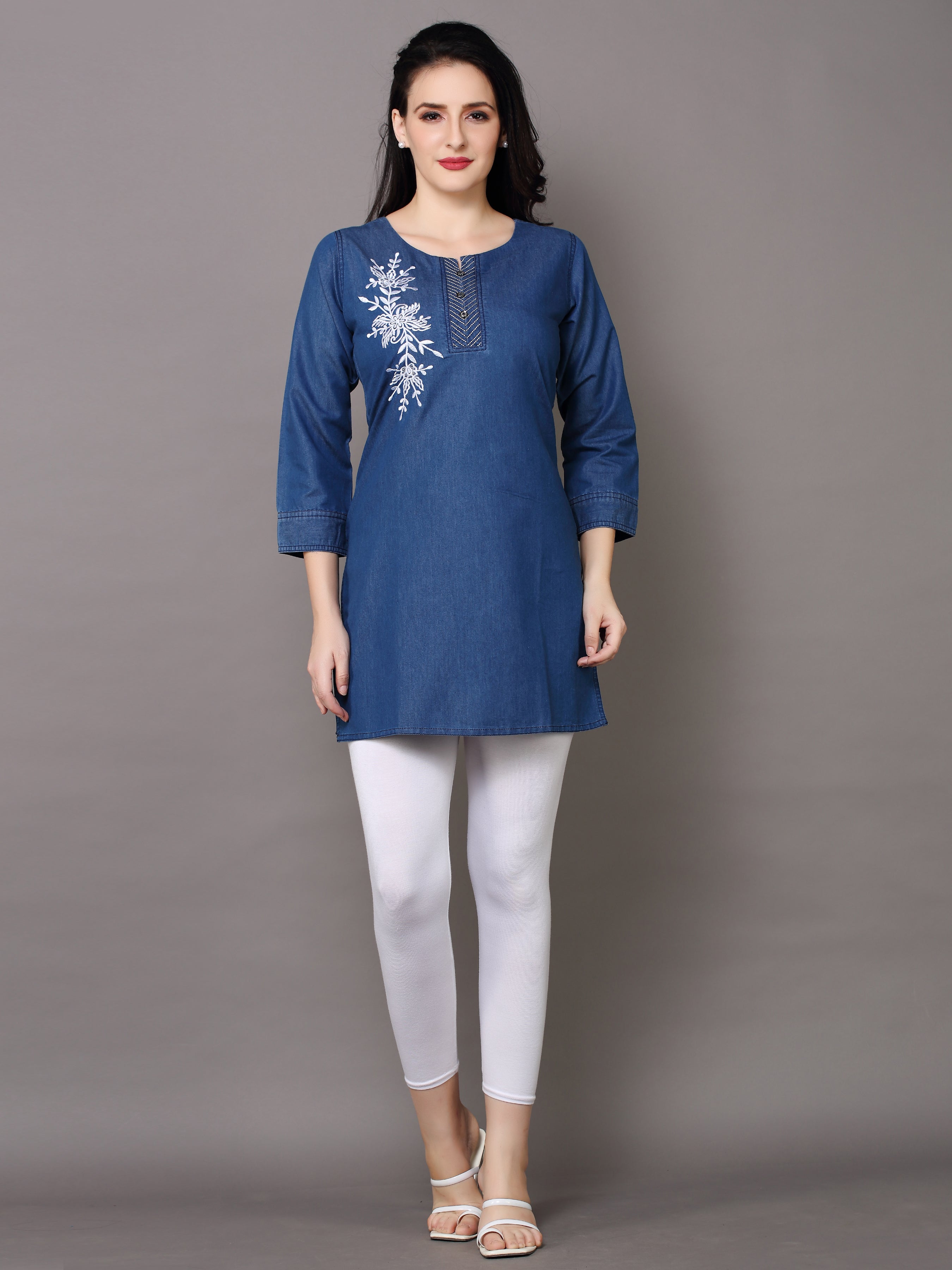 A woman modeling a blue cotton kurta with white floral embroidery on the chest, paired with white leggings and beige sandals, against a solid gray studio background.