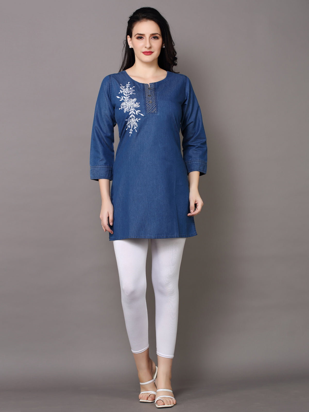 A woman modeling a blue cotton kurta with white floral embroidery on the chest, paired with white leggings and beige sandals, against a solid gray studio background.