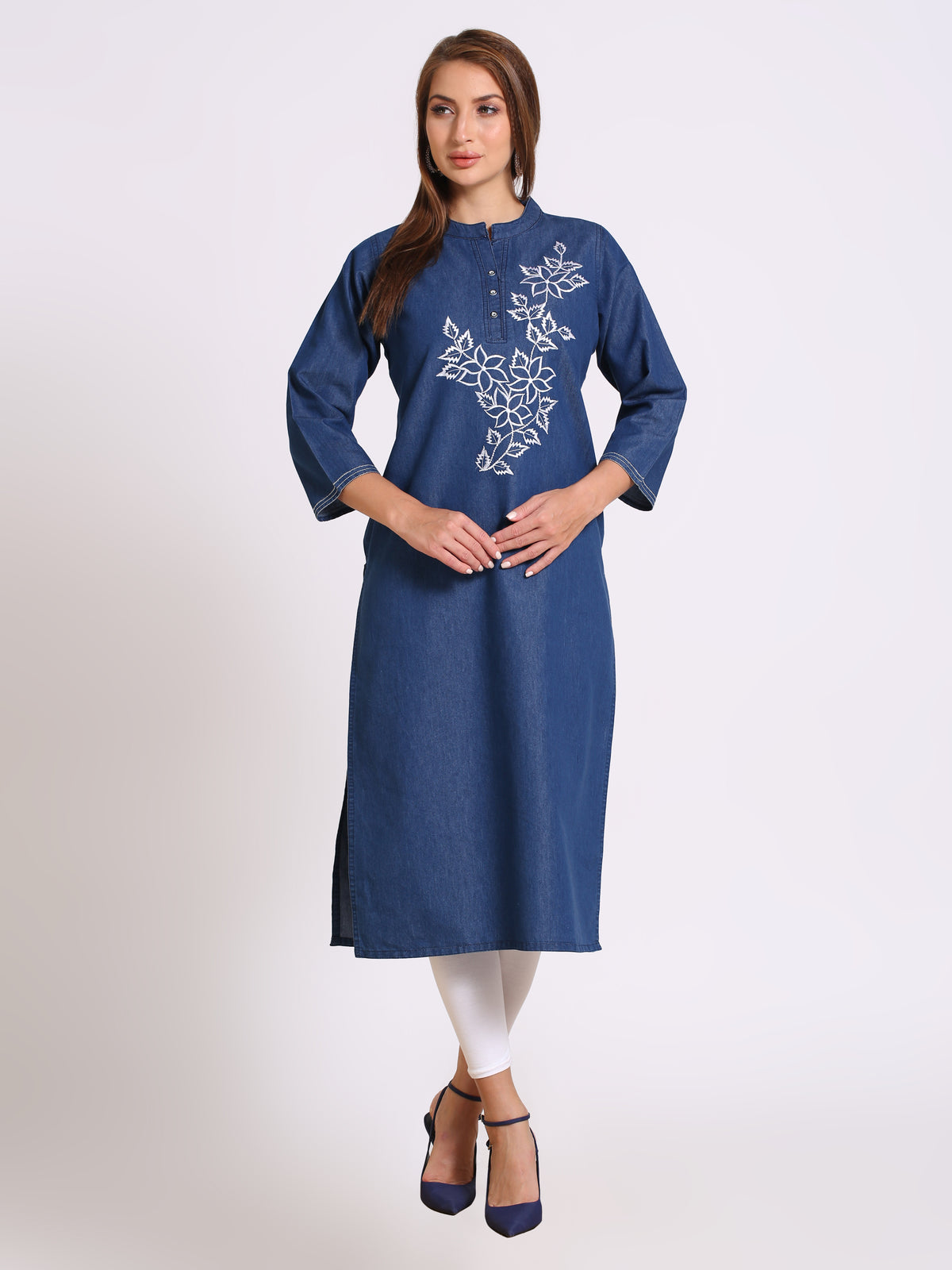 A woman modeling a blue cotton kurta with white floral embroidery on the chest, paired with white leggings and navy blue heels, against a light gray studio background.