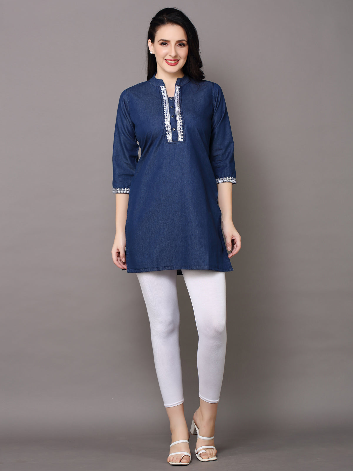 A woman modeling a navy blue kurta with white embroidered trim on the neckline and cuffs, paired with white leggings and white heeled sandals, against a solid gray studio background.