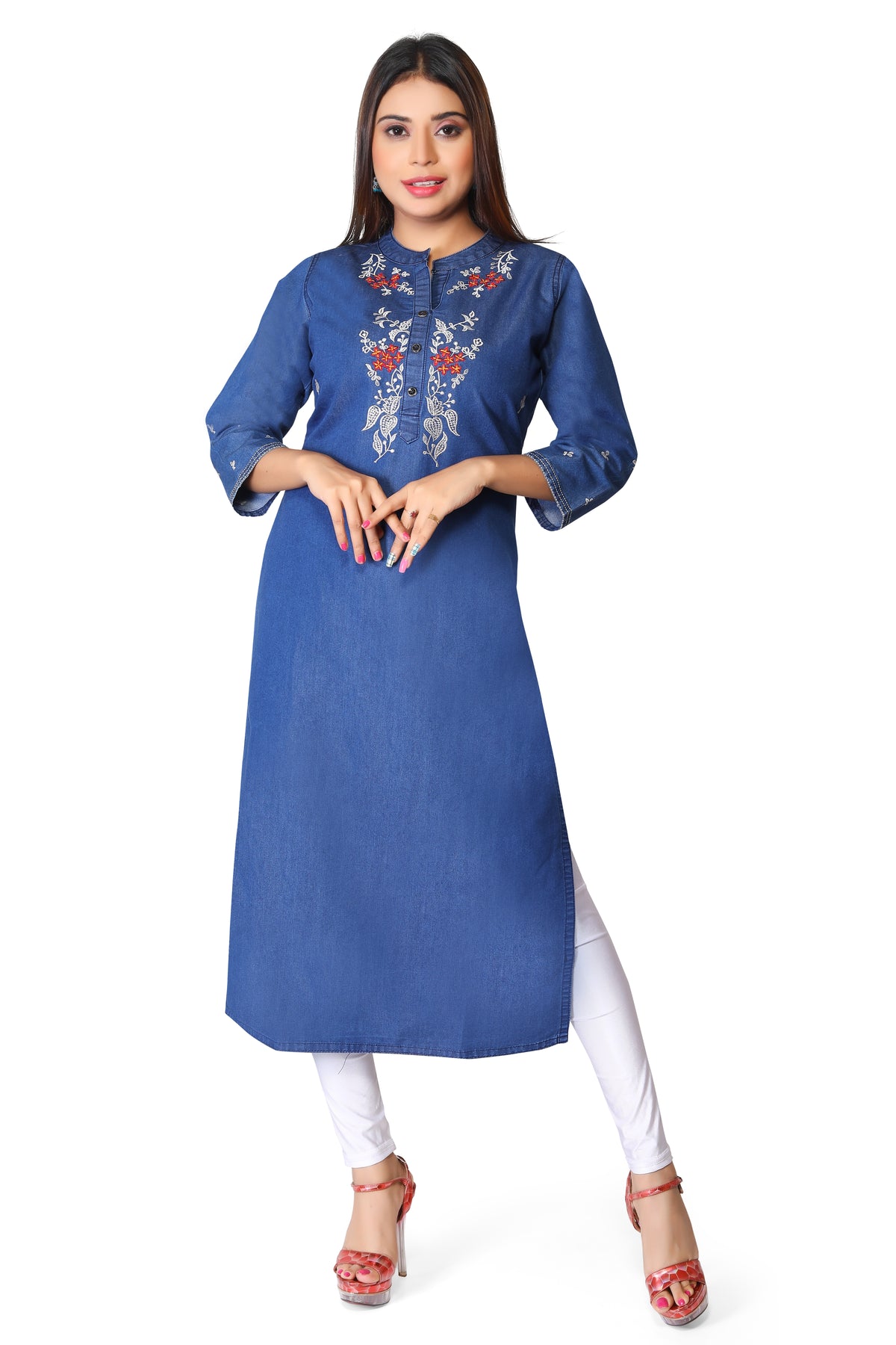 A woman modeling a blue denim-style A-line kurta with colorful floral embroidery on the chest, paired with white leggings and red high-heeled sandals, against a plain white background.