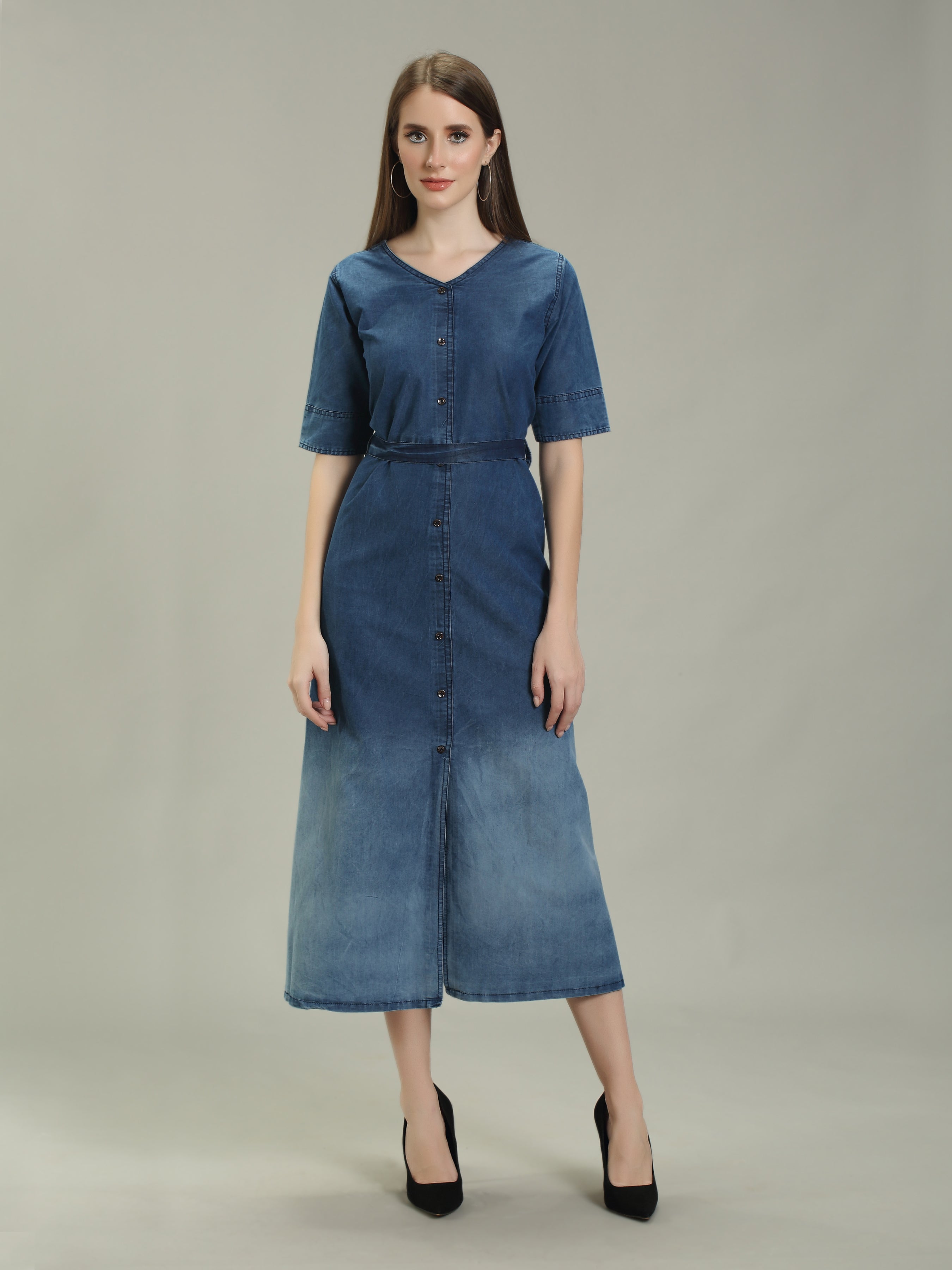 A woman modeling a short-sleeved blue denim midi dress with a button-down front and a matching waist tie, paired with black heels, against a neutral gray studio background.